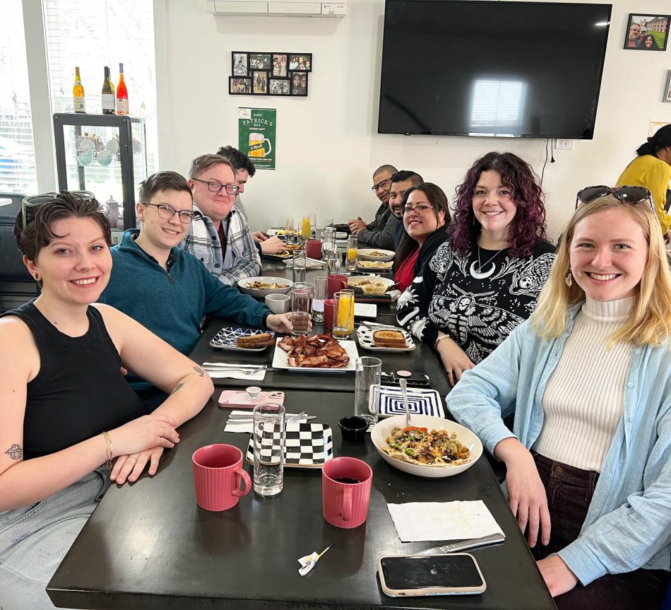 Cheerful group of diverse friends enjoying a monthly brunch at Rehana's, hosted by FAHASS' Pride Center in Fredericksburg, VA, with a spread of breakfast dishes on the table.