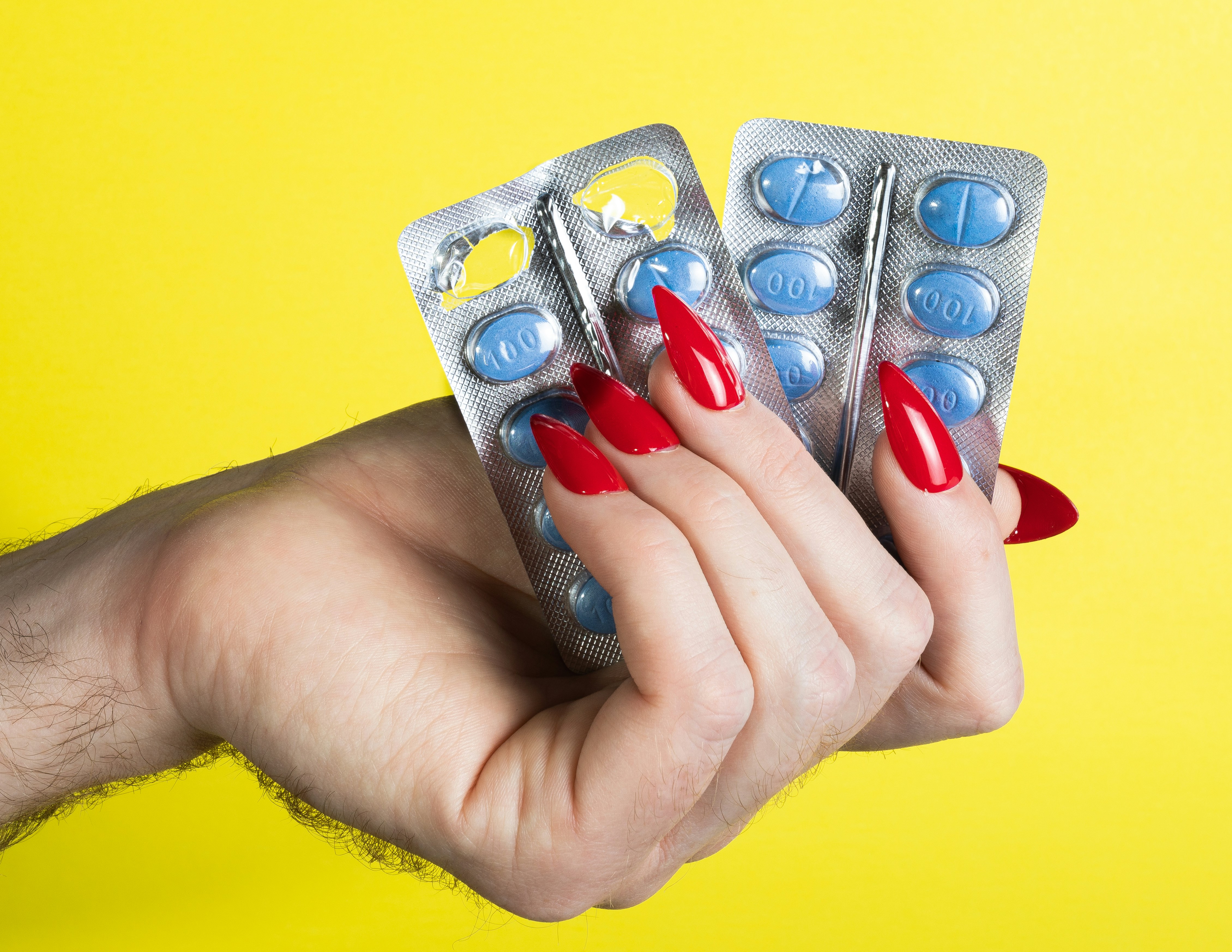 A hand holding blister packs of pills with red nails against a yellow background, symbolizing the availability of PrEP at Mosaic Care Center for HIV and STI prevention.