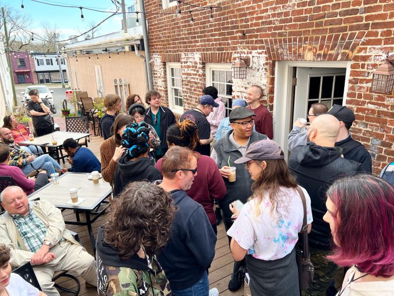 Lively gathering at the Queer Connection Coffee Social, hosted by FAHASS' Pride Center and Curitiba Art Cafe in Fredericksburg, VA, with diverse individuals chatting on an outdoor patio.