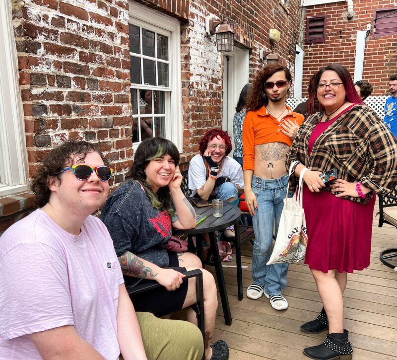 Lively gathering at the Queer Connection Coffee Social, hosted by FAHASS' Pride Center and Curitiba Art Cafe in Fredericksburg, VA, with diverse individuals on an outdoor patio.