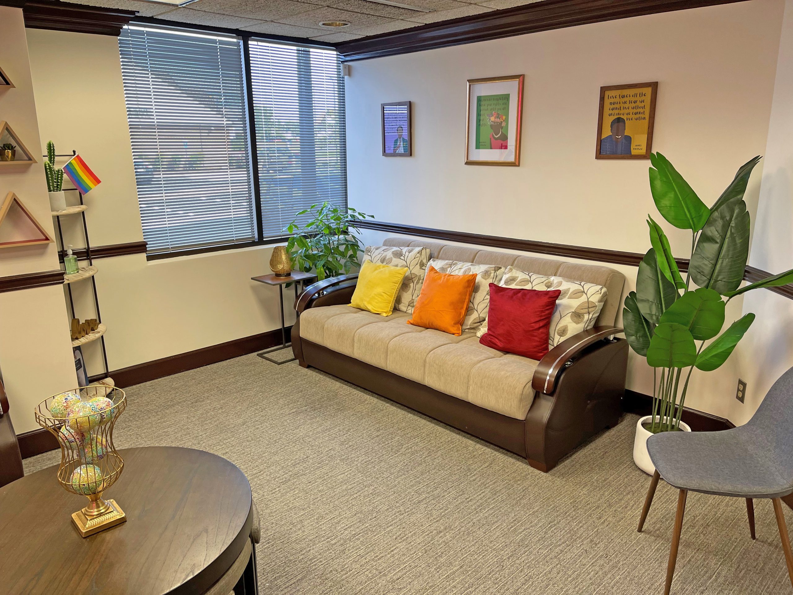 A cozy and welcoming room with a sofa, colorful pillows, plants, and a small Pride flag on a shelf, representing the safe and inclusive environment at Pride Center FXBG.